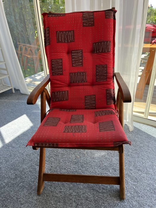 Träfåtölj med röd stoppad dyna med abstrakt mönster, placerad i ett soligt rum.