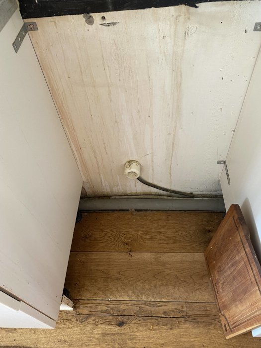 Utsnitt av ett hörn i ett rum med trägolv, oputsad vägg och synliga rör.