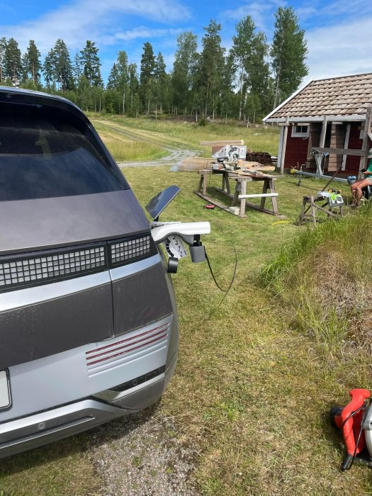 Modern elbil laddas på landsbygden med laddkabel, äldre hus och virke i bakgrunden.