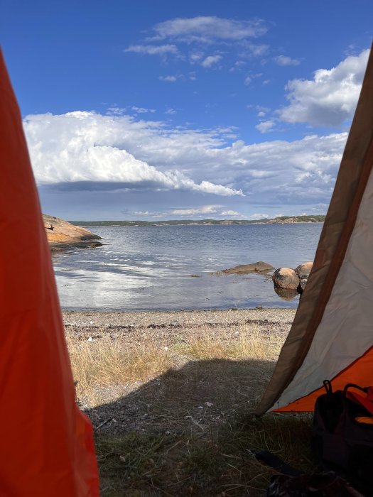 Utsikt från tältöppning mot lugn sjö och klippor under blå himmel med moln.