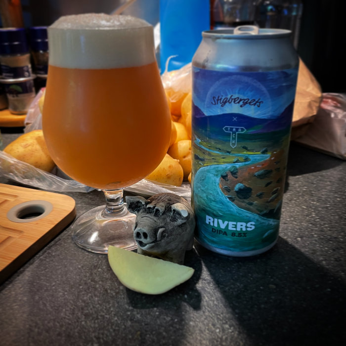 Ölglas fyllt med grumlig öl, ölburk med etikett "Rivers DIPA", ostbit och träskulptur av gris.