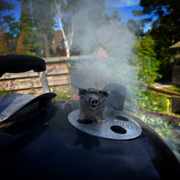 Rök stiger från en grill med en metallgrisdekoration på locket under soligt väder.