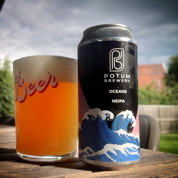 Ölburk med texten "Potum Brewery OCEANS NEIPA" och ett halvfyllt glas på en träyta utomhus.