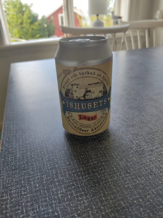 En ölburk märkt "ISHUSETS Lager" på ett bord med suddig bakgrund av ett fönster med utsikt över ett rött hus.