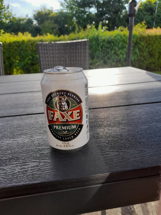 En burk Faxe Premium lageröl på ett utomhusbord med stolar och grönska i bakgrunden.