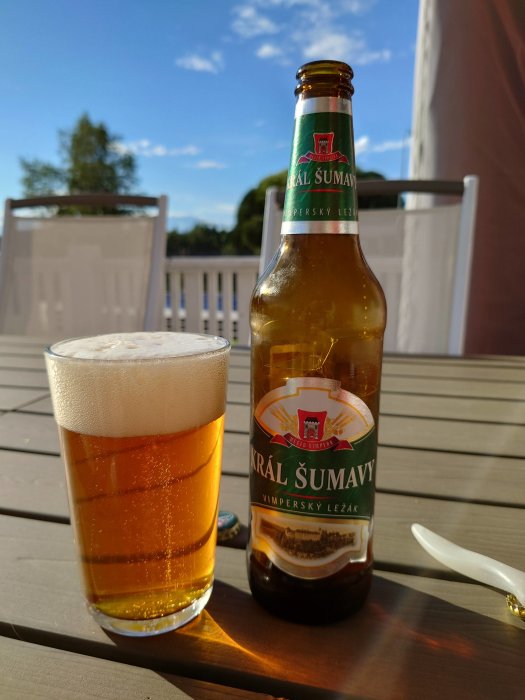 Ölflaska och glas med öl på ett träbord utomhus, med soligt landskap i bakgrunden.