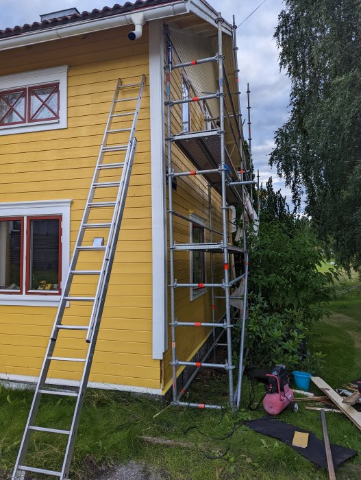 Montering av knutbrädor på gult hus med ställning och stege, verktyg på marken.