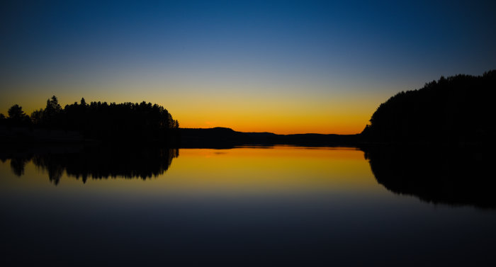 Solnedgång över en stilla sjö med en spegelbild av himlen och silhuetter av träd och berg i bakgrunden.