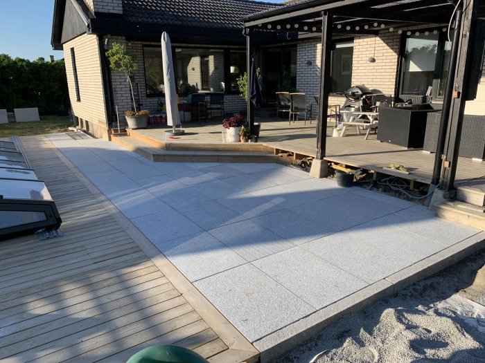 Renoverad uteplats med gråa granitplattor vid trätrall framför hus med veranda och trädgårdsmöbler.