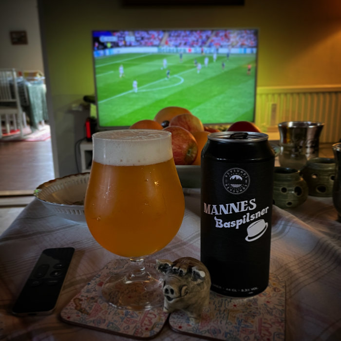 Ett glas öl framför en TV som visar fotboll, bredvid en burk Märkes Baspilsner och en fjärrkontroll.