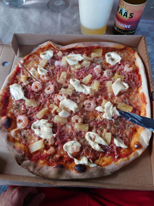 Hawaiipizza med räkor, ananas och majonnäs, lätt strödd med svartpeppar, serverad i en kartong.