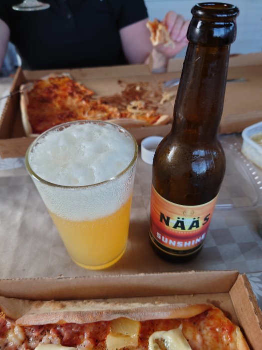 Ett glas öl bredvid en ölflaska märkt 'NÄÄS SUNSHINE', kartonger med pizza i bakgrunden.