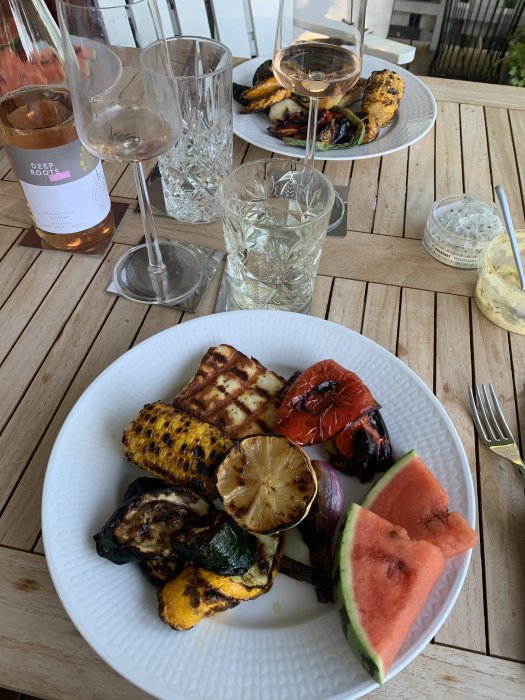 Grillade grönsaker och vattenmelon på tallrik med rosévin på träbord.