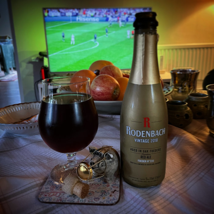 Ett glas Red Ale och en flaska Rodenbach Vintage 2018 på ett bord med en fotbollsmatch på TV i bakgrunden.