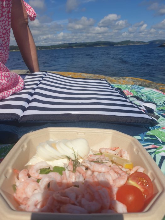 Räkmacka i take-away-förpackning på en randig kudde vid havet, person i bakgrunden.