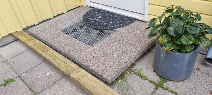 En granittrappa med inbyggd gallerdurk och en prydnadsmatta vid en entré, stödd av två trävärdar, med krukväxt intill.