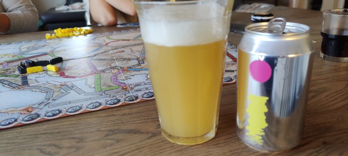 Ett glas öl framför en burk Omnipollo Fatamorgana med Ticket To Ride-brädspel i bakgrunden.