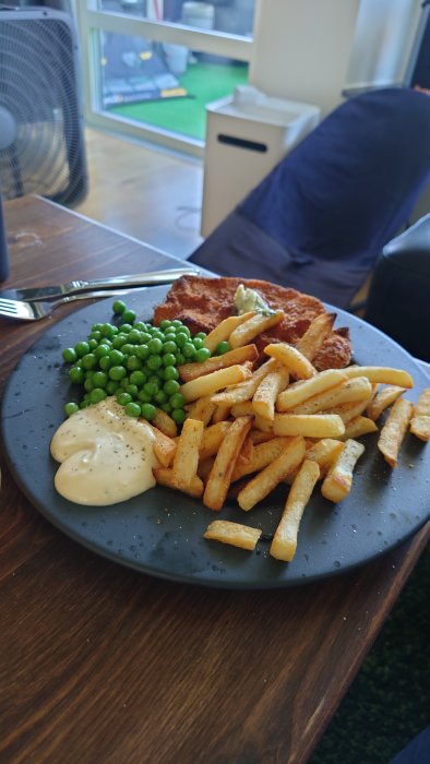 En tallrik med schnitzel, pommes frites, ärtor och bearnaisesås på ett träbord.