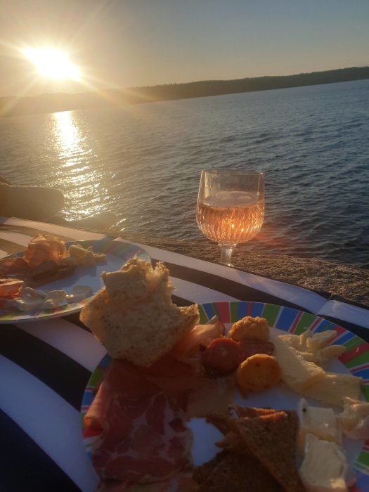 Solnedgång vid havet med ett glas vin och en tallrik med ost, charkuterier och bröd på klipporna.