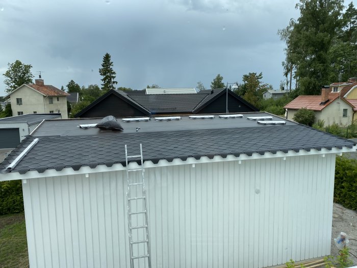 Nytt svart tak på ett garage med en steg som leder upp till taket, omgivet av villabebyggelse under en mulen himmel.