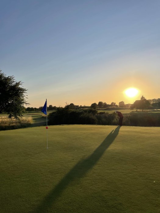 Solnedgång över golfbana med flagga och spelares skugga på greenen.