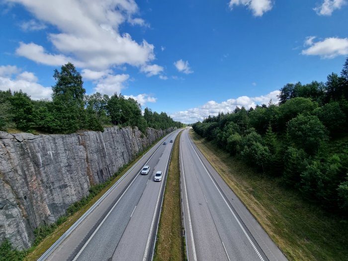 Vy över en motorväg med bilar mellan klippväggar och gröna träd under en blå himmel med vita moln.