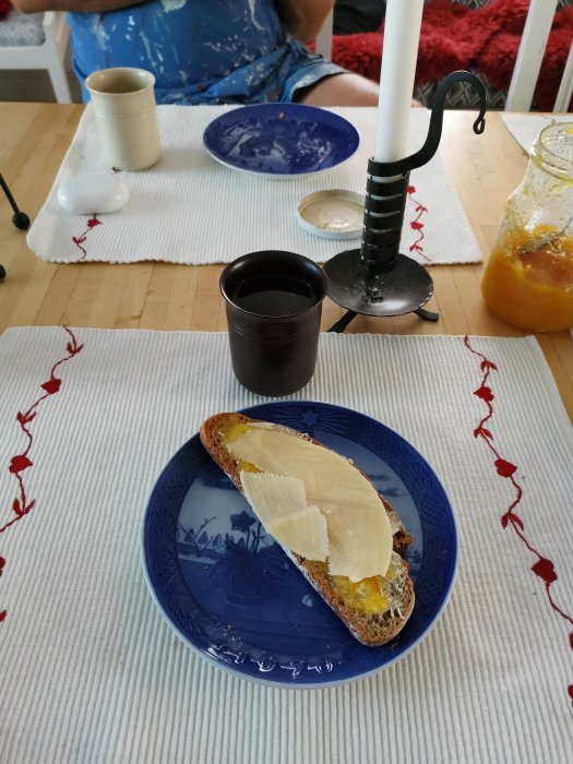 Skiva av hembakt surdegsbröd med apelsinmarmelad och cheddar på blå tallrik, burk med marmelad, ljus och koppar.