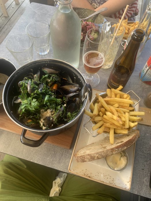 En gryta med musslor och persilja, pommes frites, bröd med sås och öl på restaurangbord.