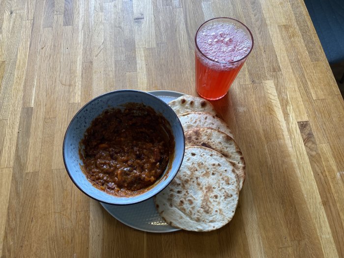 Chiligryta i skål med bröd och ett glas röd vinbärssaft på träbord.