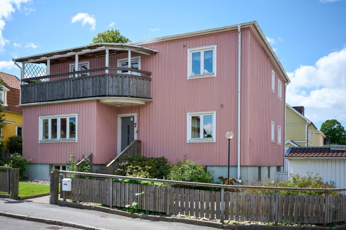 Tvåvånings rosa hus med balkong och trästaket på en solig dag.