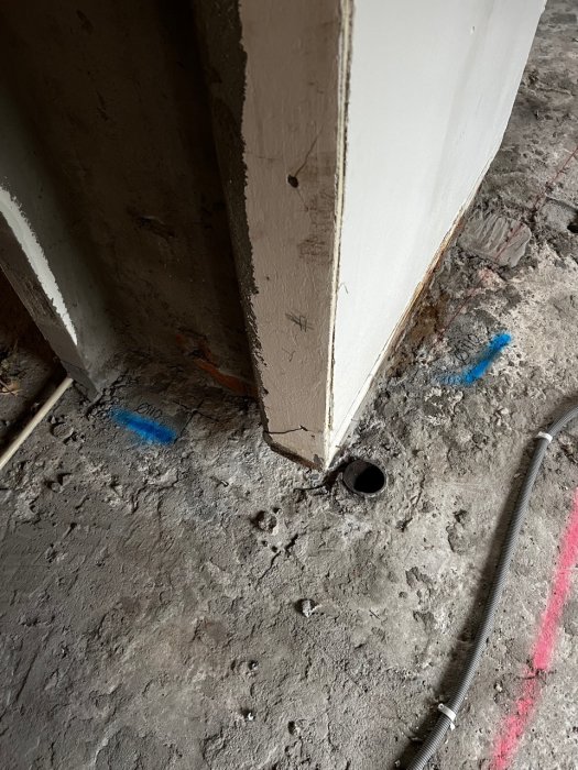 Hörn av ett betonggolv med markerade punkter och ett borrat hål vid en vägg, kabel och rör syns.