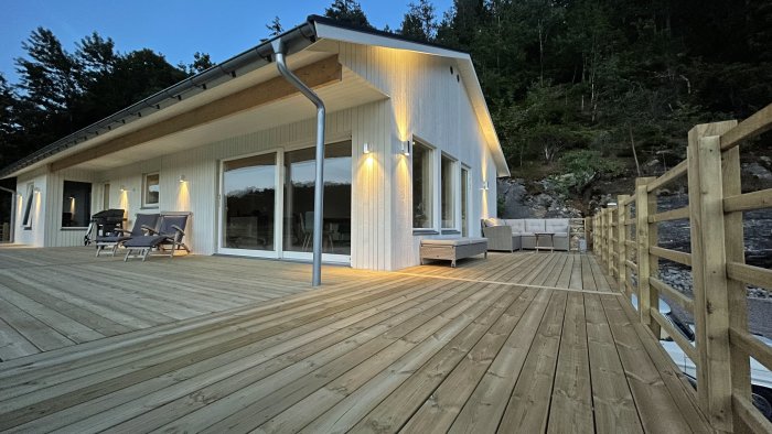 Trätrall på en modern villa med stora fönster och lounge-möbler i skymning med belysning och skog i bakgrunden.