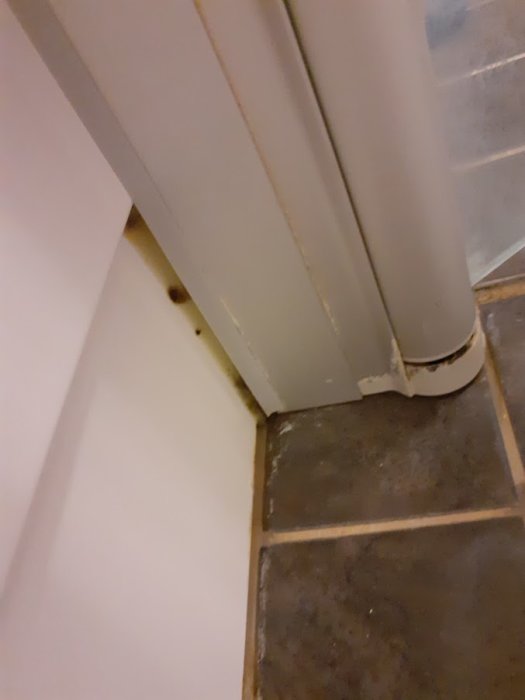 Skadad fotlist och bortglidande täckning vid rörgenomföring i hörn mellan vägg och golv.
