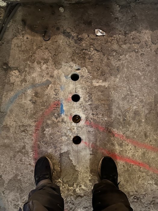 Ett betonggolv med uppmärkningar och tre borrade hål för avloppsrör, och personens skor syns i förgrunden.