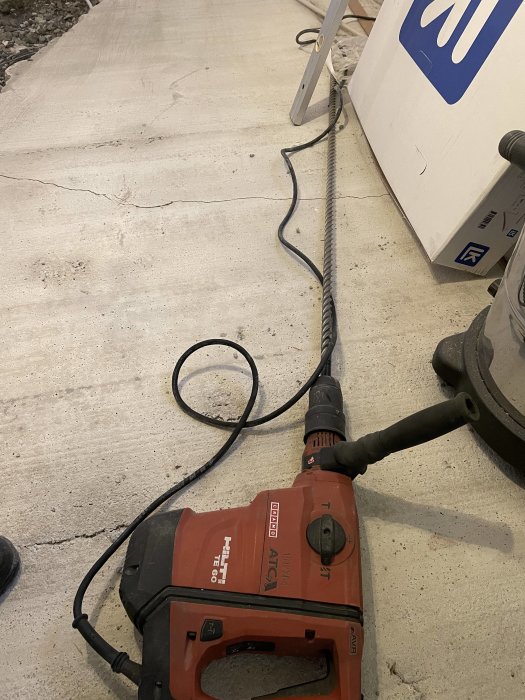 En hilti slagborr på ett betonggolv bredvid en uppbruten platta och borrspån, vilket visar renoveringsarbetet.