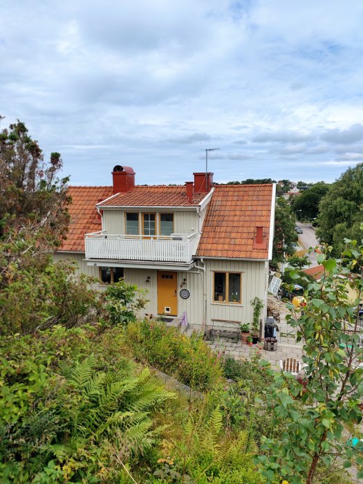 Vy över ett traditionellt hus med tegeltak, balkong och gul ytterdörr omgiven av grönska.