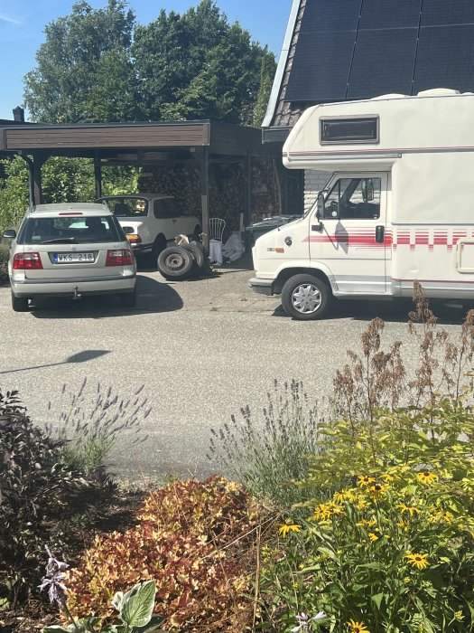 Husvagn och personbil parkerade på en uppfart bredvid en vedbod och blomstrande trädgård.