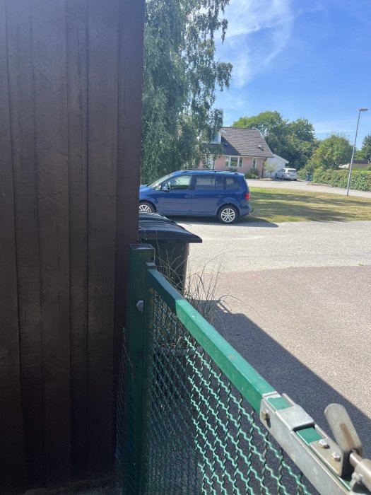 Blå bil parkerad framför ett hus sett från bakom en brun träplank och grön nätstaket på en solig dag.