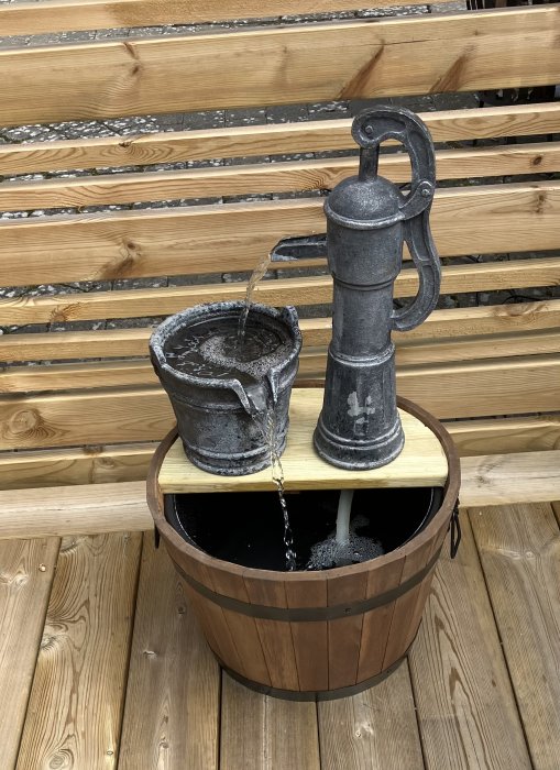 Trähink med vatten och en handpump i plast som pumpar vatten i en behållare, placerade på en träterrass.