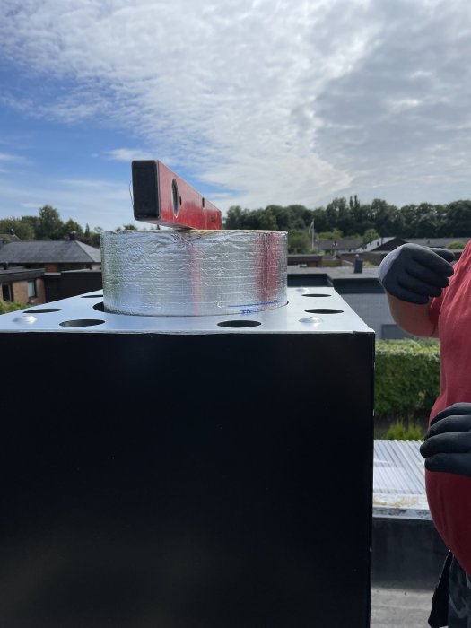 Vattenpass och rulle med packtejp på toppen av en svart ventilationsenhet, utomhus med blå himmel.