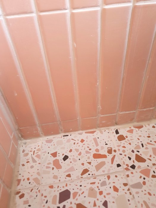 Hörnan av ett badrum med rosa kakelväggar och terrazzomönstrat golv i neutrala toner.