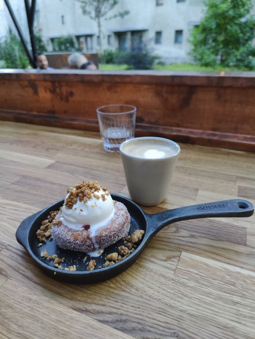 Efterrätt bestående av en donut med glass och smulor i en liten gjutjärnspanna bredvid en kopp kaffe på ett träbord.