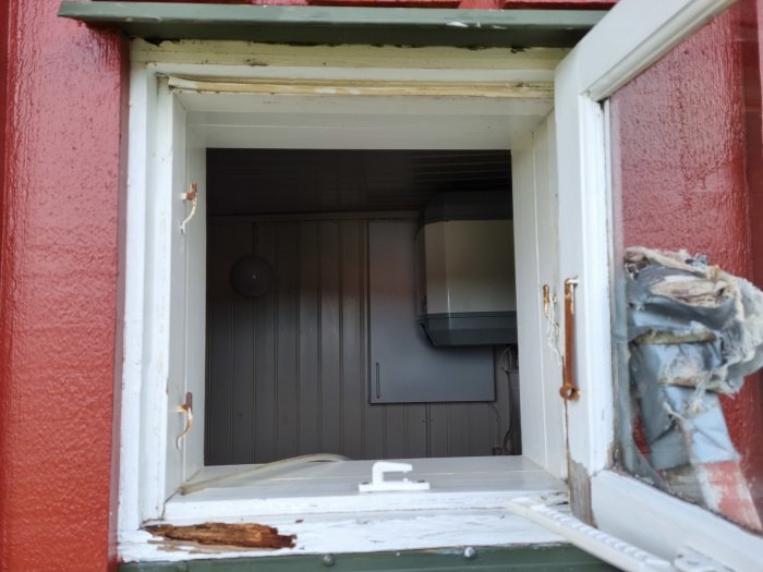 Skadat fönster med flagnande färg och en titt in i ett mörklagt rum med synlig trapp och lampett.