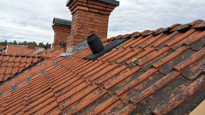 Röd tegelpannor på ett tak med skorsten och ventilationstrumma, omgivet av annan bebyggelse.