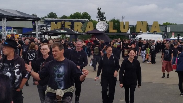 Människor i olika åldrar och stilar promenerar vid en festival med "M'ERA LUNA" skylt i bakgrunden.