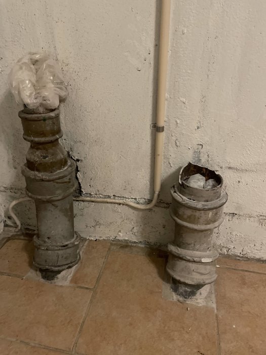 Gamla avloppsrör i betong med en öppen del och en tillsluten med plastpåse, intill en vit vägg.