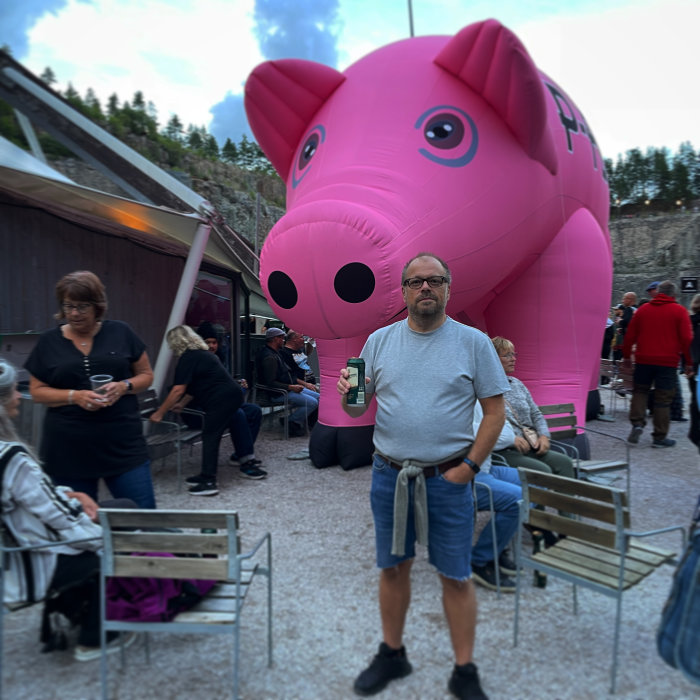 Man står framför en stor uppblåsbar rosa gris i ett utomhusevenemang med människor runtomkring.