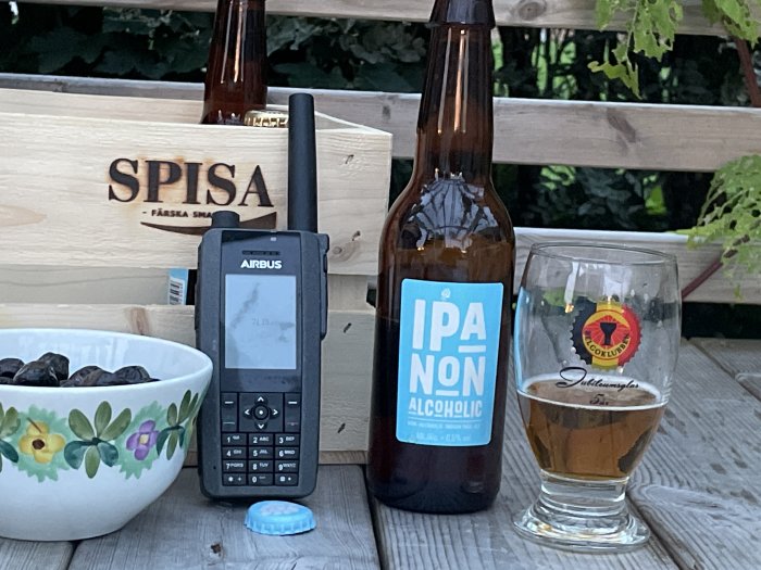 Utomhusfikabord med alkoholfri IPA-flaska, walkie-talkie, skål med oliver och ölglas mot trälåda märkt "SPISE".