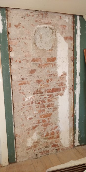 En delvis avskalad vägg där tegelstenar och rester av vit puts syns, omgiven av träpanel och listverk.