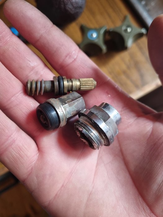 En hand som håller en isärplockad mekanisk del, möjligtvis en kran eller ventil, med fler komponenter i bakgrunden.